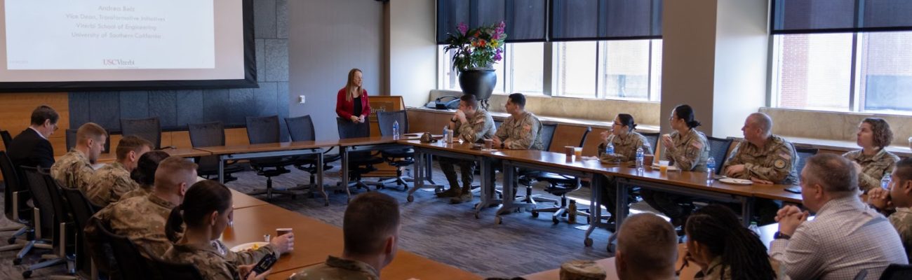 People in military uniforms sit at a square shaped desk while a women stands and presents. The powerpoint slide reads "Welcome to USSF Polaris Awardees November 18, 2022"